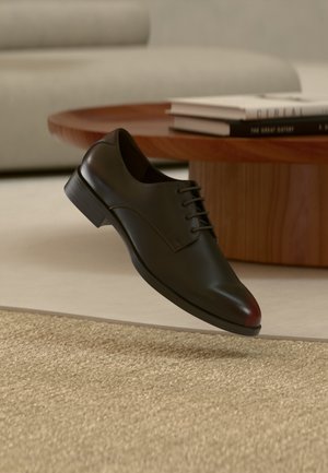 Chaussure habillée en cuir noir avec lacets flottant au-dessus d'un tapis beige texturé, table ronde en bois avec des livres empilés en arrière-plan.