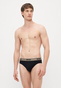 Juodos kelnaitės su rudos spalvos liemeniu, ant kurio puikuojasi „BOSS“ logotipas. Audinys atrodo lygus ir tamprus, skirtas patogumui.