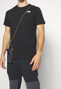 Black cotton t-shirt with a small white logo, paired with dark pants featuring grey panels and a small black pouch hanging from a braided strap.