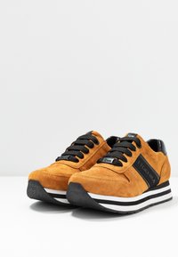 Zapatillas de ante naranja con cordones y rayas negras, que presentan una puntera redondeada y suela de goma negra texturizada. Diseñadas con un estilo deportivo.