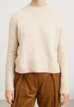 Personne portant un pull en maille côtelée crème à manches longues et un pantalon en velours côtelé marron, debout devant un fond clair uni.