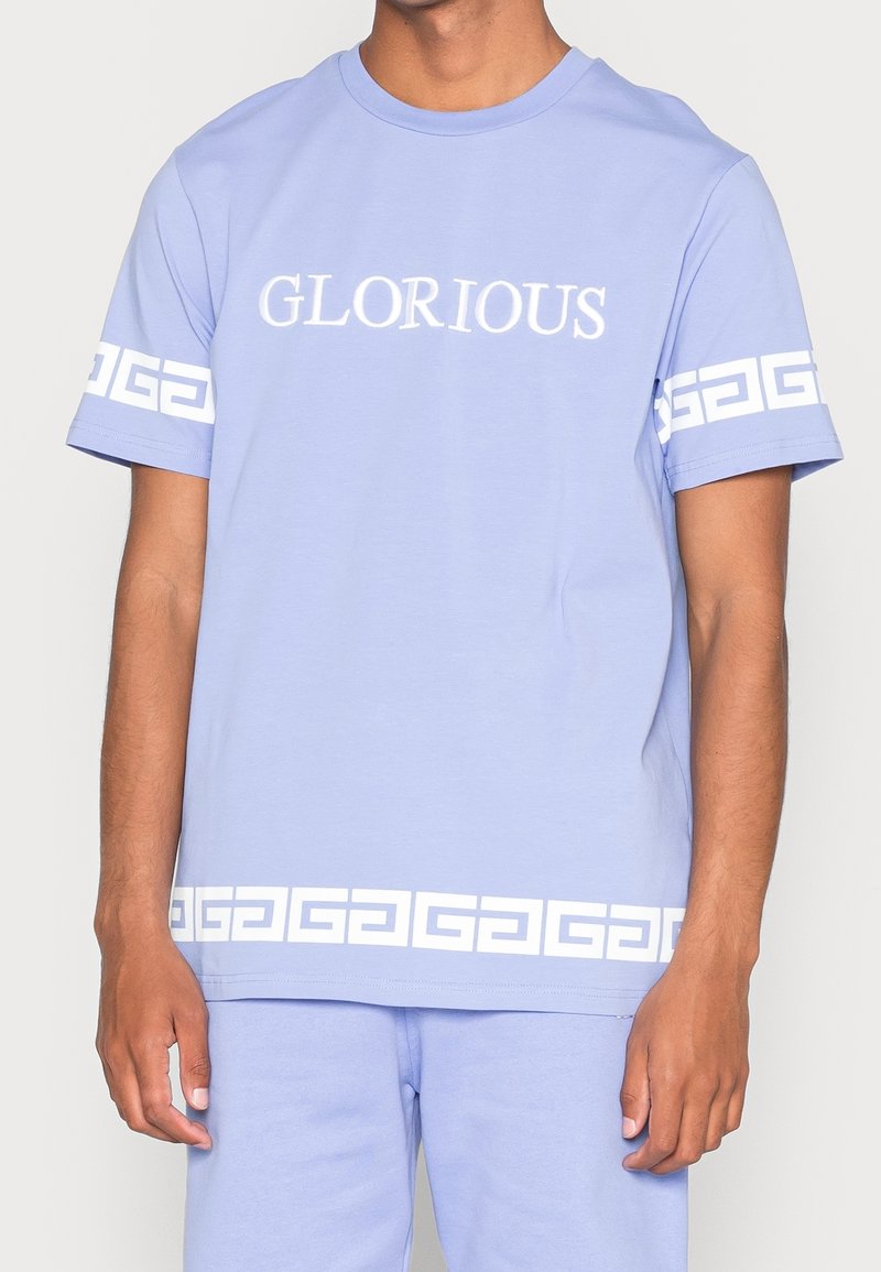 Camiseta de algodón de color azul claro con "GLORIOSO" impreso en blanco, y un patrón geométrico blanco en las mangas y el dobladillo inferior.