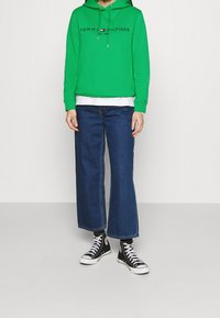 Grön hoodie med dragsko, varumärkeslogotyp i svart och röd. Vidbeniga blå jeans och svarta högre sneakers med vita sulor.