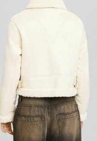 Kremowa kurtka ze skóry owczej o skróconym kroju, z teksturowanym futrzanym kołnierzem, przeszyciami i paskiem w talii.