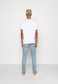 T-shirt en coton blanc, manches courtes, associé à un jean denim bleu clair. Le modèle est vu de dos, portant des baskets jaunes. Design simple.
