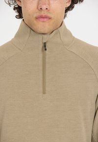 Beige halv-zip trøje lavet af blødt, tekstureret stof. Den har en høj krave og en sort lynlås med en trækklap. Letvægtsdesign.