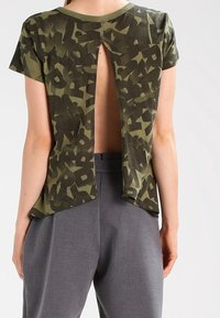 Olivově zelené tričko s abstraktním černým vzorem, s klíčkovým výstřihem na zádech a krátkými rukávy. Spárováno se šedými kalhotami s vysokým pasem.