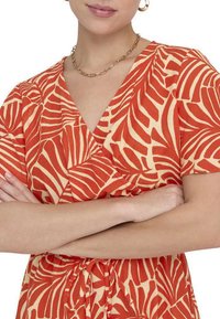 Robe-rappe rouge et crème avec motif de feuilles, manches courtes, décolleté en V et détail à la taille à nouer, fabriquée à partir d'un tissu léger, texture douce.