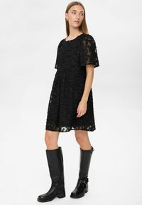 Rochie neagră cu o textură din dantelă florală, mâneci scurte și talie ajustată. Asortată cu cizme din piele neagră până la genunchi.
