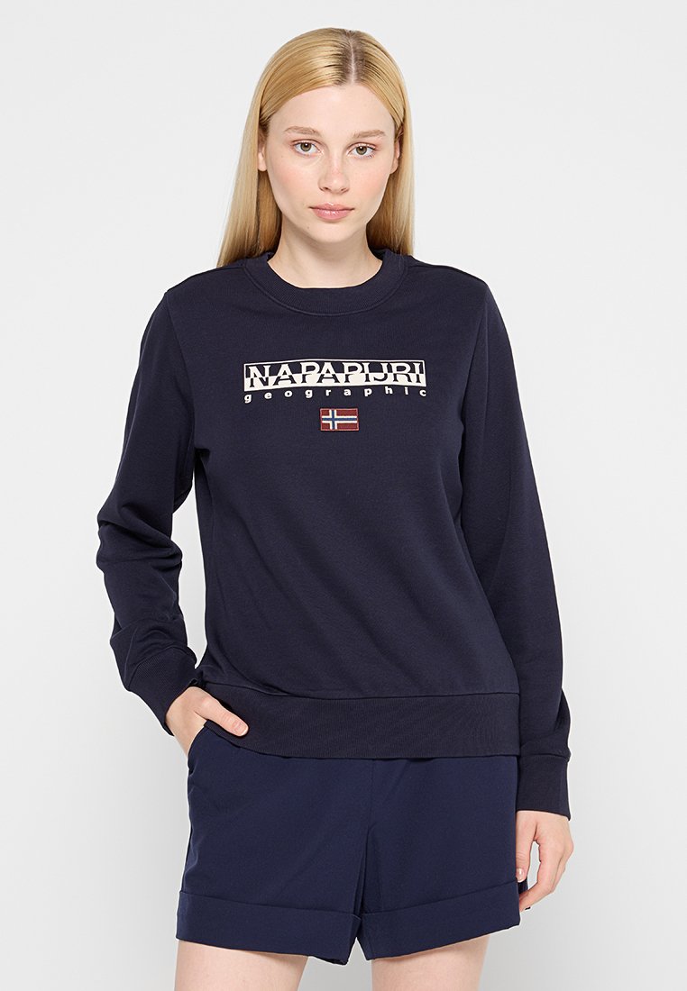 Giovane donna con lunghi capelli biondi che indossa una felpa color navy con la scritta "NAPAPIJRI geographic" e la bandiera norvegese, abbinata a pantaloni corti navy.