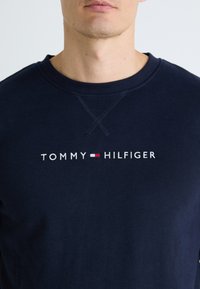 Mies, jolla on päällään tummansininen huppari, jossa on "Tommy Hilfiger" -teksti ja pieni punainen, valkoinen ja sininen lippulogo rinnassa, yksinkertaista taustaa vasten.