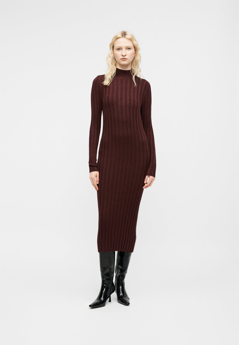 Femme aux cheveux blonds clairs portant une longue robe-pull côtelée bordeaux et des bottes noires montantes à talons, debout devant un fond gris uni.