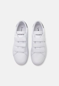 Fehér Lacoste sportcipők három tépőzárral, kerek orral, sima szintetikus anyagból, logóval a nyelvén és fekete díszítéssel a sarkán.