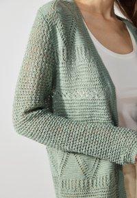 Ανοιχτό πράσινο πλεκτό cardigan με ανοιχτή ύφανση, που διαθέτει V-λαιμόκοψη και μακριά μανίκια, ελαφρώς τοποθετημένο πάνω από ένα λευκό μπλουζάκι.