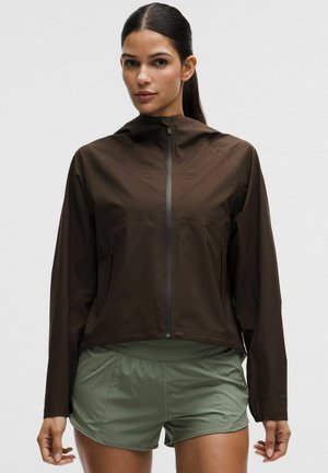 Kobieta o ciemnych włosach związanych z tyłu nosi brązową kurtkę zapinaną na zamek z kapturem i zielone sportowe szorty, stoi na tle jasnego, jednolitego tła.