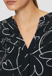Blouse noire avec des motifs de cœurs blancs et du texte manuscrit. Design en V, texture lisse et plis subtils au niveau de l'encolure.