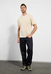 Camiseta corta de manga corta color beige claro con finas rayas verticales, combinada con jeans de color azul oscuro y zapatillas verdes oliva con patrón.