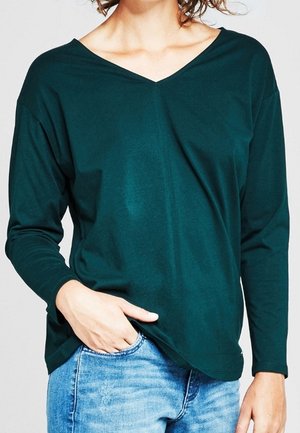 Personne portant un t-shirt à manches longues vert foncé avec un col en V et un jean bleu clair, une main dans la poche avant.
