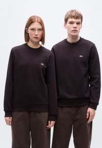 Μαύρα βαμβακερά φουτεράκια με στρογγυλή λαιμόκοψη, διακοσμημένα με μικρά πράσινα κεντημένα λογότυπα. Συνδυάζονται με σκουρόχρωμα καφέ παντελόνια, λείο ύφασμα, άνετη εφαρμογή.