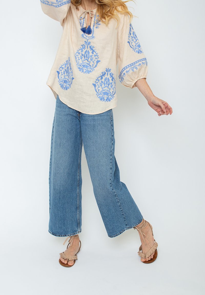 Femme portant un chemisier crème avec broderies florales bleues, un jean bleu à jambes larges, et des sandales beige à volants sur fond blanc.