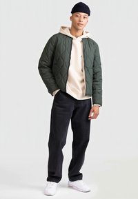 Prešívaná zelená bomber bunda cez béžovú mikinu, kombinovaná s čiernymi džínsami a bielymi teniskami. Bunda má okrúhly golier a zips.