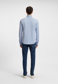 Camisa de manga larga azul claro con cuello clásico, textura suave y dobladillo curvado, combinada con jeans de mezclilla oscura y zapatos de color claro.