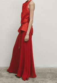 Blouse rouge sans manches avec un ourlet asymétrique, associée à une jupe rouge fluide. Le tissu semble texturé et léger, avec des embellissements subtils.