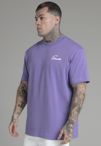 Lavendel farvet bomulds t-shirt med korte ærmer og rund hals, præget af hvid kursiv branding på venstre bryst. Designet til en afslappet pasform.