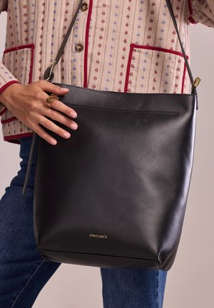 Sac en cuir noir avec une texture lisse, des détails de logo doré et un design simple. Il dispose d'une longue bandoulière et d'une fermeture à glissière.