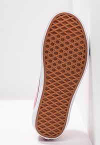 Semelle d'une basket avec semelle extérieure en caoutchouc marron à motif gaufré et semelle intermédiaire blanche, positionnée verticalement contre une surface blanche.