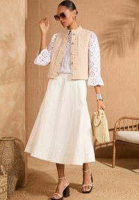 Beige sleeveless vest, white eyelet blouse, white skirt, and block-heeled sandals. Textured fabric, braiding on vest, patterned sleeves.