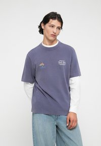 Violeta kokvilnas T-kreklis ar apaļu kaklu, īsām piedurknēm, ar izšūtu zivju dizainu un tekstu. Uzvilkts virs balta garām piedurknēm krekla.