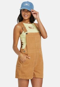 Salopette in corduroy marrone con una vestibilità rilassata, dotata di spalline regolabili, tasche laterali, indossata sopra un top a coste verde chiaro.