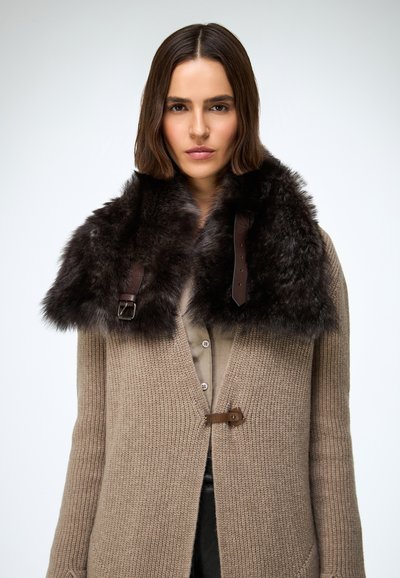 Beež kudumiskašmiiriga kardigan pruunide karvade kraega. Krae on kaunistatud pruunide nahkrihmade ja pannaldega, mis loob tekstuurse kontrasti.