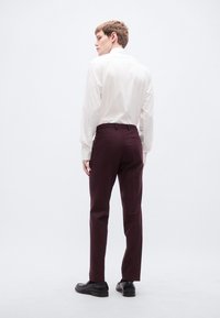 Λευκό πουκάμισο με μακριά μανίκια, φορεμένο με σκούρο μπουρντό παντελόνι, εμφανιζόμενο από πίσω, με μαύρα παπούτσια και μια ομαλή, προσαρμοσμένη εφαρμογή.