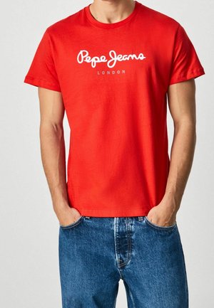 Mężczyzna w czerwonej koszulce Pepe Jeans London i niebieskich jeansach, ręce w kieszeniach, stoi na jednolitym jasnym tle.