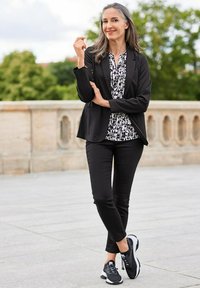 Zwarte blazer over een wit en zwart patroon blouse, gecombineerd met zwarte broek en zwarte sportschoenen, staand buiten met groen in de achtergrond.