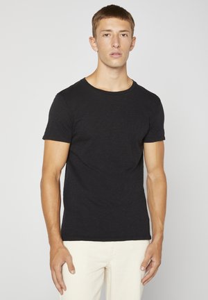 T-shirt nera a maniche corte realizzata in un tessuto morbido e texturizzato, con scollatura a girocollo e una silhouette aderente, abbinata a pantaloni chiari.