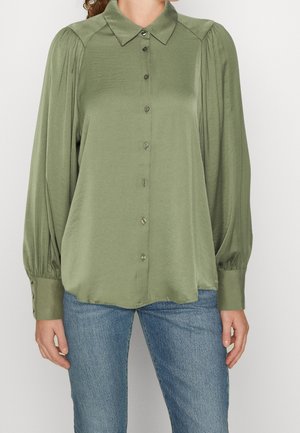 Mujer con blusa de botones de manga larga verde oliva con hombros plisados y vaqueros azules, de pie frente a un fondo claro y liso.