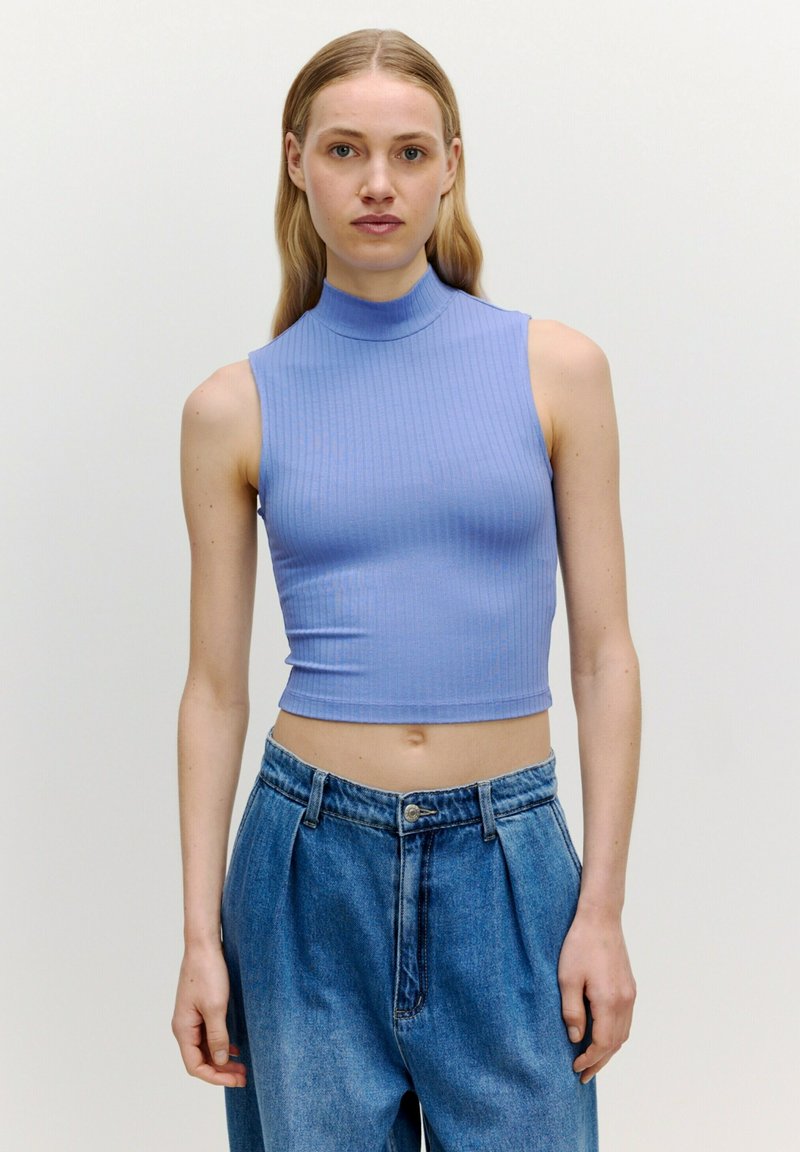 Lichtblauw geribd mouwloos topje met hoge hals, gecombineerd met een losse spijkerbroek in blauw, voorzien van een knoopsluiting en voorplooien.