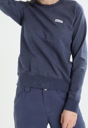 Person trägt einen dunkelblauen Patagonia-Sweatshirt und passende Hose, mit einem Arm erhoben und dem anderen seitlich ruhend.