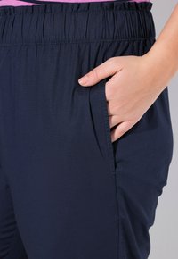 Marinblå byxor med elastisk midja och sidofickor. Tyget ser slätt ut med en lätt textur.