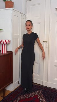 Γυναίκα με τατουάζ φορά μακρύ μαύρο φόρεμα και κόκκινο κραγιόν, στέκεται σε γωνία κοντά σε λευκές πόρτες και ξύλινα έπιπλα με διακοσμητικό μπολ.