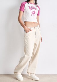 Camiseta blanca recortada con mangas rosas y gráfico "HUGO". Pantalones anchos crema con costuras azules y bolsillos laterales. Zapatillas blancas.