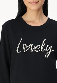 Sweatshirt noir avec l'inscription "lovely" embellie de accents blancs en forme de perles ; encolure ronde et texture douce.