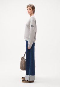 Grå hettegenser med ribbestrikkede mansjetter, vide blå jeans med slitt kant, og brune sandaler. Modellen holder en grå totebag.