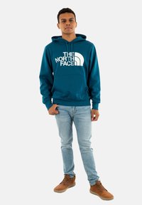 Sudadera verde con logotipo blanco, bolsillo canguro, puños y dobladillo acanalados. Combinada con jeans azul claros y botas de tobillo marrones con cordones.