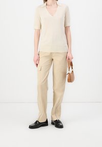 Maglione beige a maniche corte con scollo a V, abbinato a pantaloni cargo beige con tasche laterali e dettagli con zip. Scarpe eleganti nere e una borsa marrone.