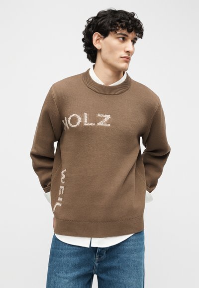 Νέος άνδρας με σγουρά μαλλιά, φορώντας καφέ πλεκτό πουλόβερ με λευκό κείμενο, λευκό πουκάμισο από κάτω και μπλε τζιν, με τα χέρια πίσω από την πλάτη.