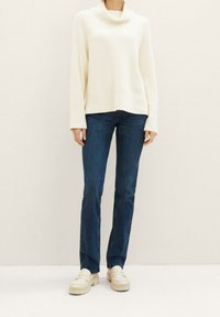 Personne portant un pull à col roulé côtelé crème, un jean droit bleu foncé et des mocassins épais écrus, debout sur un sol clair.